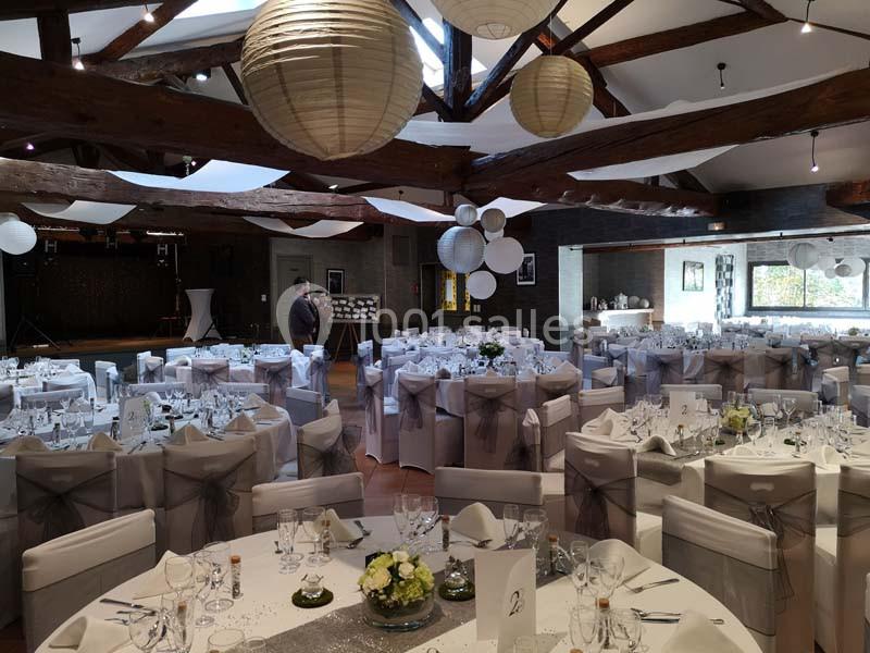 Salle de réception décorée avec des tables rondes, nappes blanches, chaises ornées de rubans gris et lanternes suspendues.