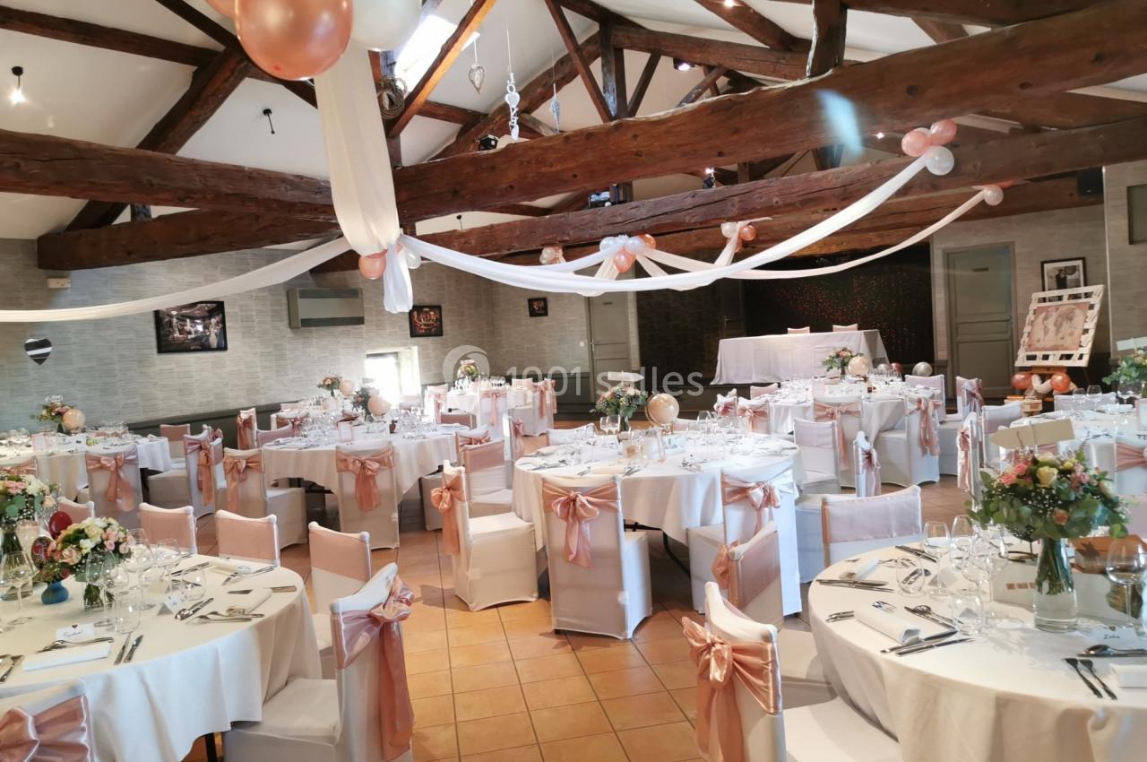 Salle de réception décorée pour un événement, avec tables dressées, nappes blanches et ornements roses.