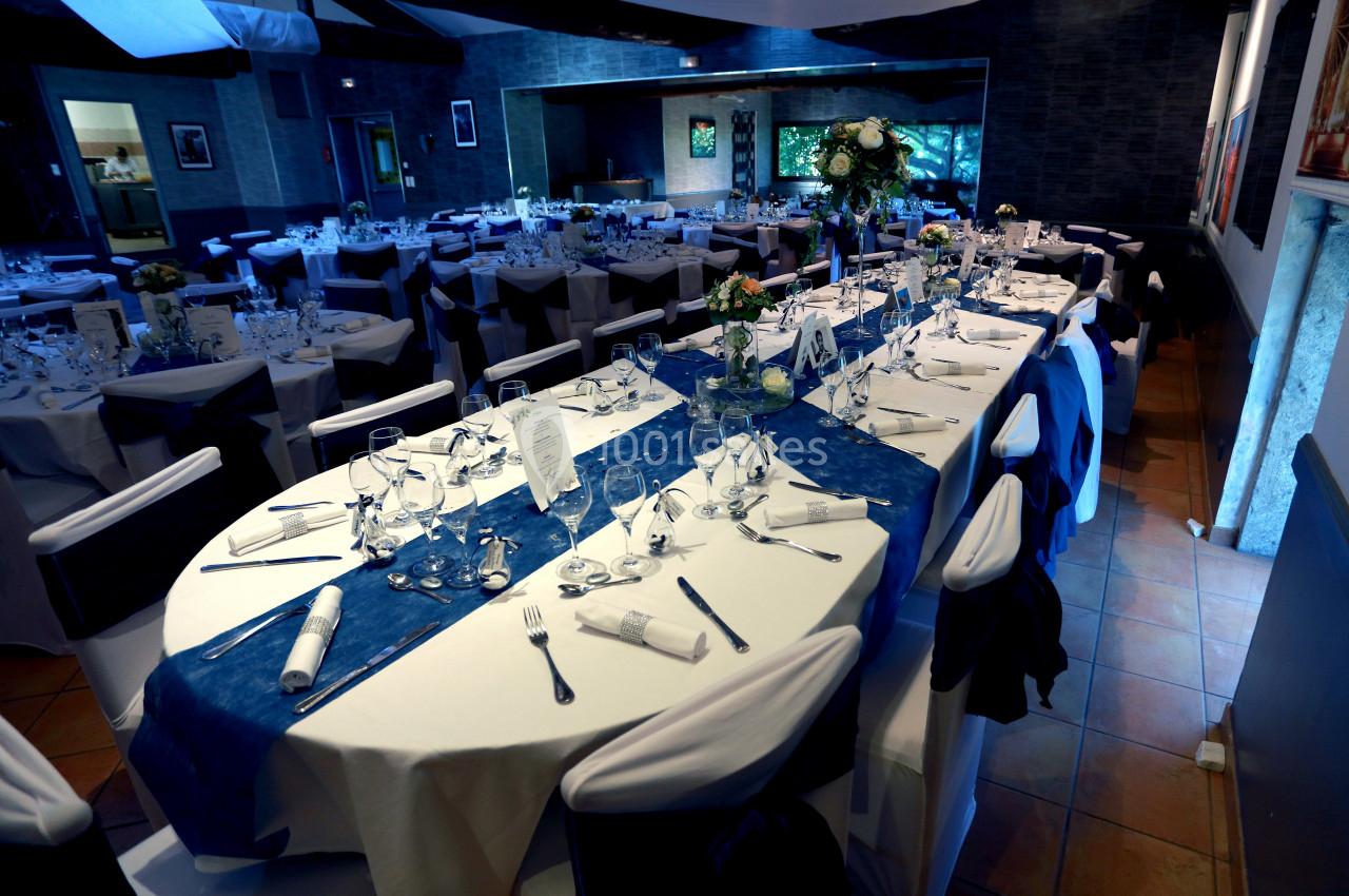 Salle de réception décorée avec des tables dressées, nappes blanches et chemins de table bleus, prête pour un événement.