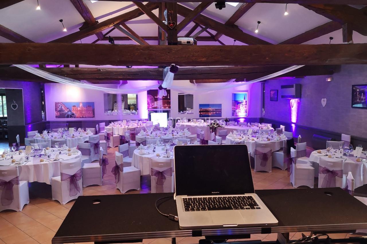 Salle de réception décorée pour un événement, avec tables dressées et éclairages violets, vue depuis une table de DJ.