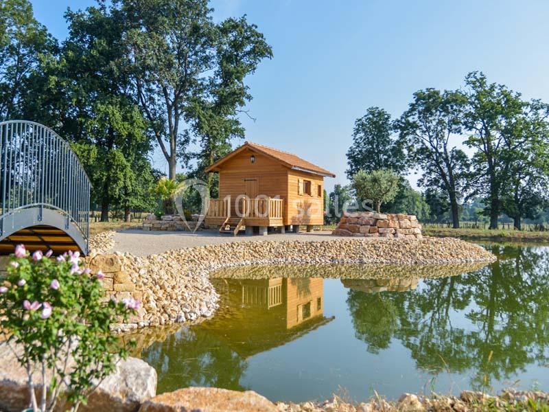 Cabane en bois au bord d'un étang entouré de végétation, avec un pont métallique et des pierres décoratives.