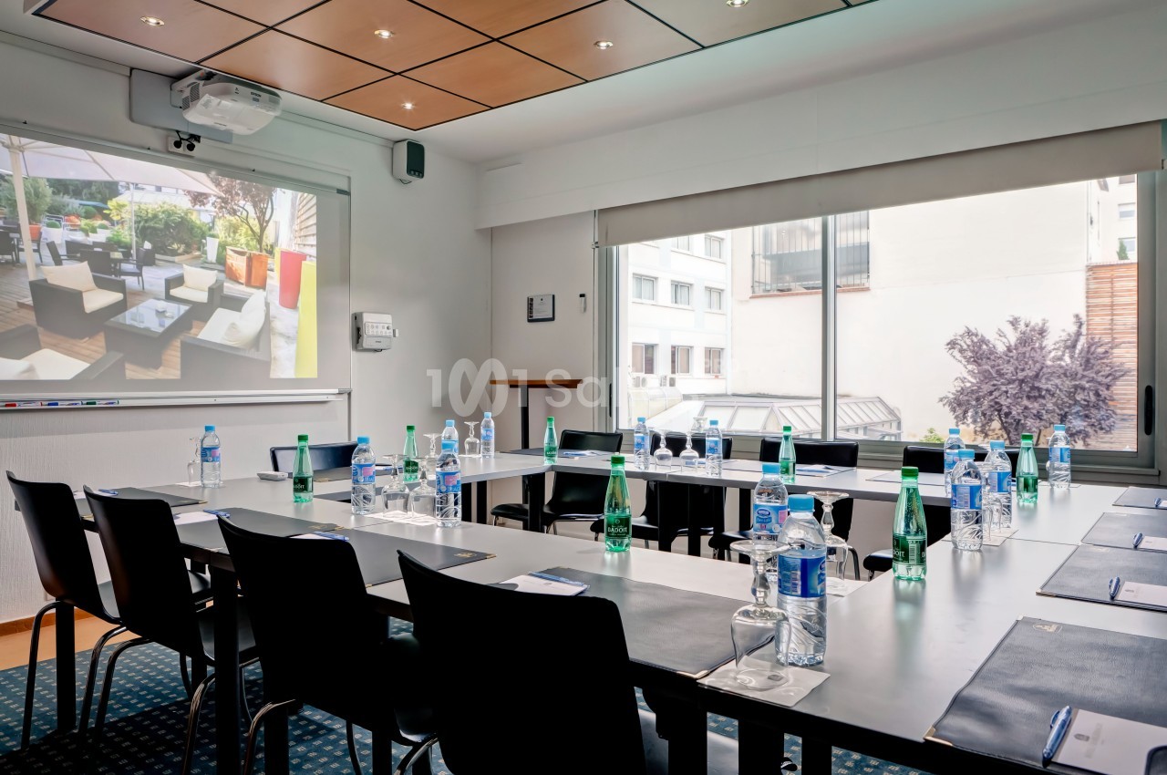 Salle de réunion lumineuse avec tables disposées en U, bouteilles d'eau, carnets et écran de projection allumé.