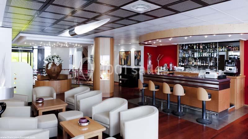 Bar moderne avec comptoir en bois, tabourets hauts, tables basses et fauteuils dans un espace lumineux et élégant.