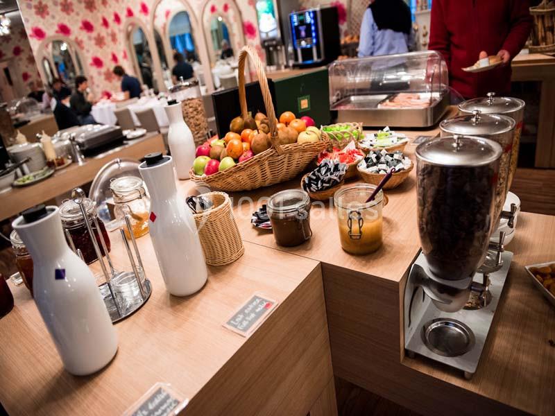 Buffet de petit-déjeuner avec fruits, céréales, boissons et viennoiseries disposés sur des tables en bois.