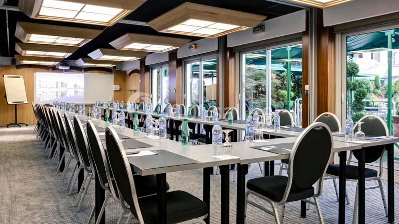 Salle de réunion lumineuse avec des tables alignées, chaises noires, bouteilles d'eau et un écran de projection.