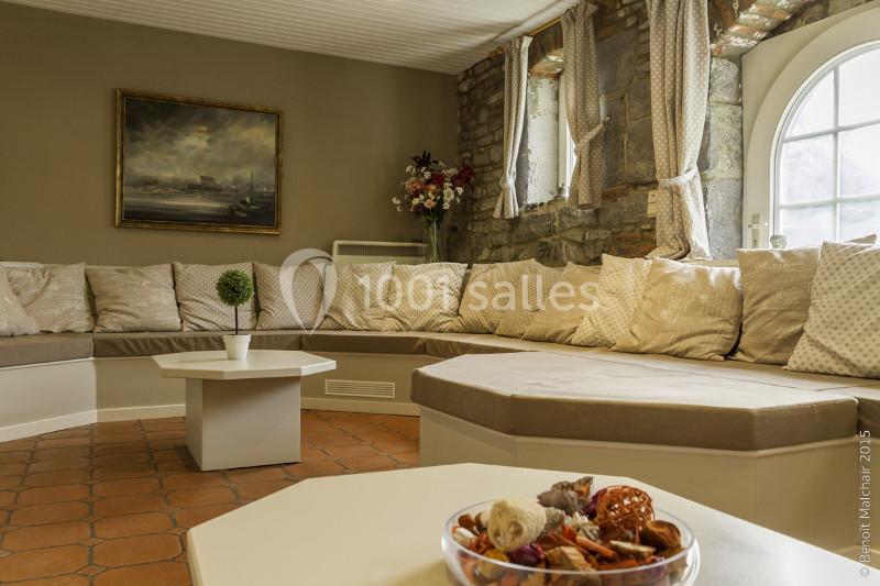 Location salle Anhée (Namur) - Ferme de l'Abbaye de Moulins #19 Salon chaleureux avec banquette d'angle beige, table basse blanche, mur en briques et fenêtre arrondie.