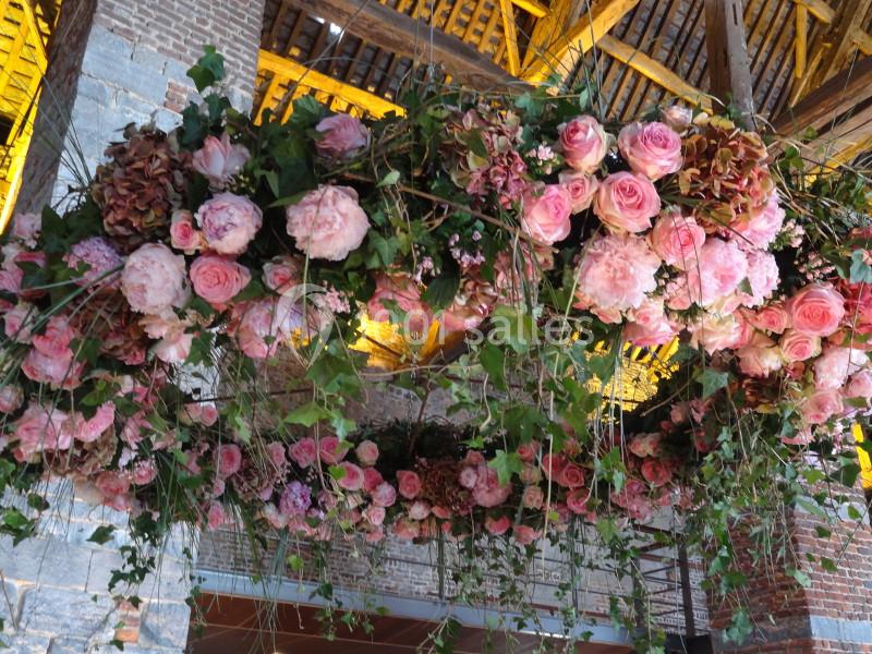 Location salle Anhée (Namur) - Ferme de l'Abbaye de Moulins #13 Guirlande suspendue de fleurs roses et feuillages dans un bâtiment en pierre et bois avec plafond en poutres apparentes.