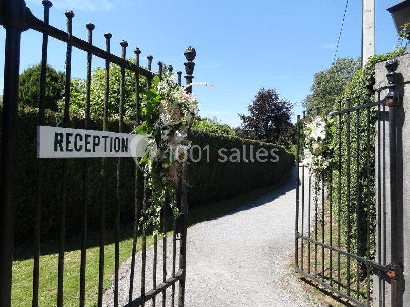 Location salle Anhée (Namur) - Ferme de l'Abbaye de Moulins #2 Portail en fer forgé ouvert, décoré de fleurs blanches, avec un panneau indiquant ’Réception’ et une allée en arrière-plan.