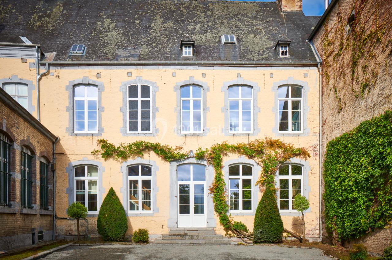 Location salle Anhée (Namur) - Ferme de l'Abbaye de Moulins #21 Façade d'un bâtiment ancien en briques jaunes avec des fenêtres blanches et des plantes grimpantes.