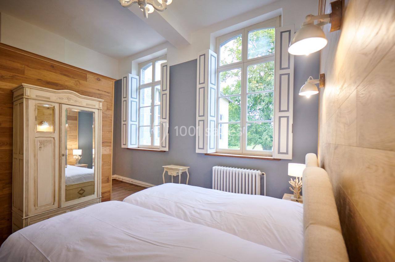 Location salle Anhée (Namur) - Ferme de l'Abbaye de Moulins #23 Chambre lumineuse avec deux lits simples, armoire ancienne, murs en bois et fenêtres donnant sur un jardin.
