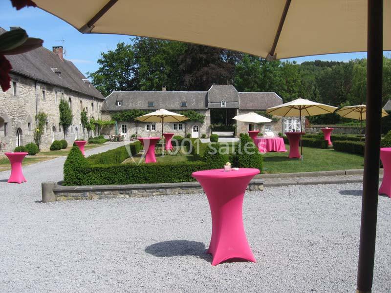 Location salle Anhée (Namur) - Ferme de l'Abbaye de Moulins #3 Cour extérieure d'un domaine avec des tables hautes roses et des parasols beiges, entourée de verdure et de bâtiments en…