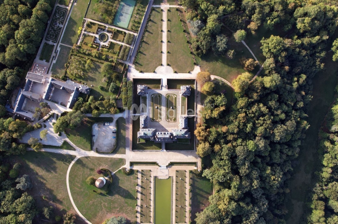 Vue aérienne d'un château entouré de jardins symétriques, d'allées et d'une forêt dense.