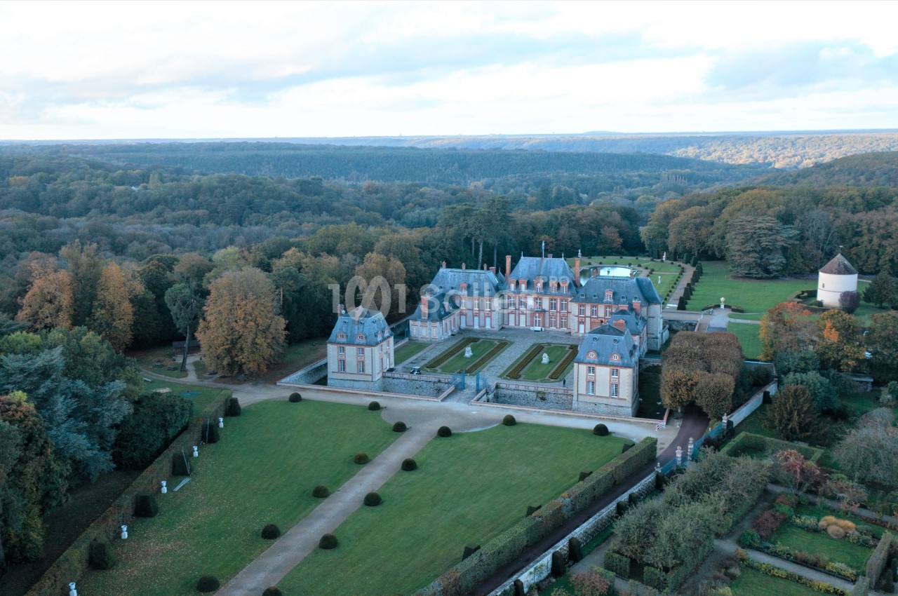 Vue aérienne d'un château entouré de jardins symétriques, d'arbres et d'une campagne boisée.