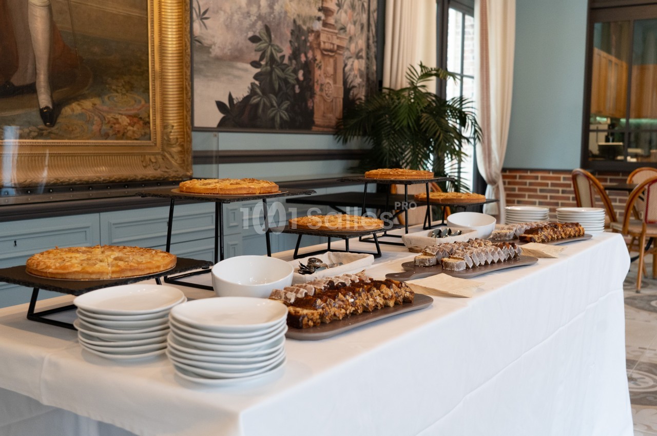 Table dressée avec des assiettes, des plats de tartes et de desserts variés, dans une salle décorée de tableaux et de…