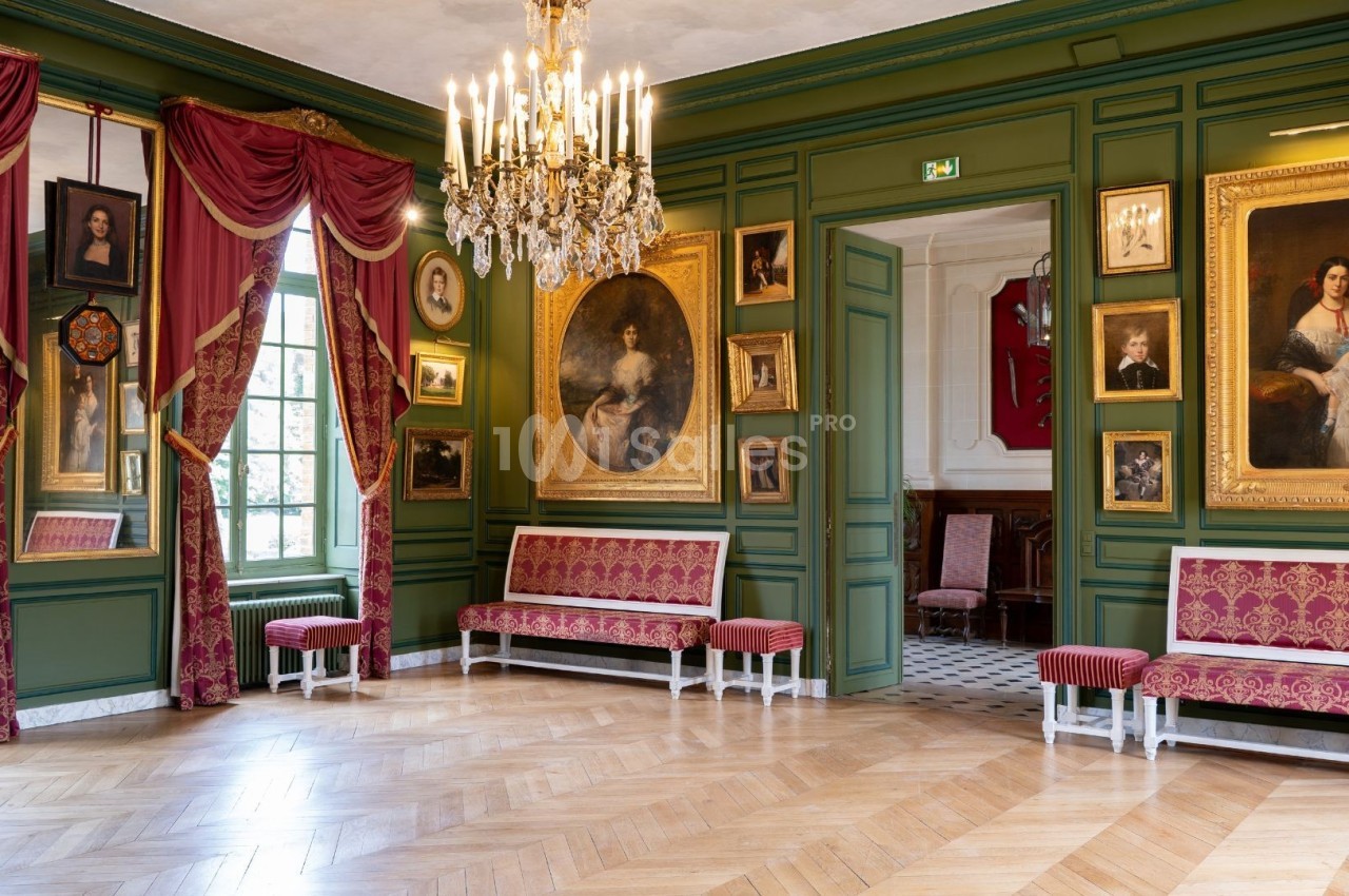 Salle élégante avec murs verts, tableaux encadrés, lustre doré, rideaux rouges et mobilier assorti.
