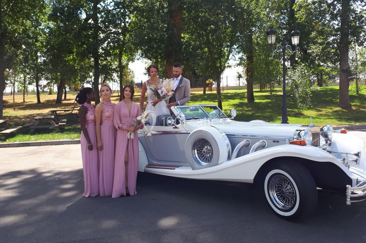 Un couple de mariés et quatre demoiselles d'honneur posent près d'une voiture blanche ancienne dans un parc.
