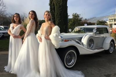 Un couple en tenue de mariage pose près d'une voiture ancienne blanche et grise devant une église en pierre.