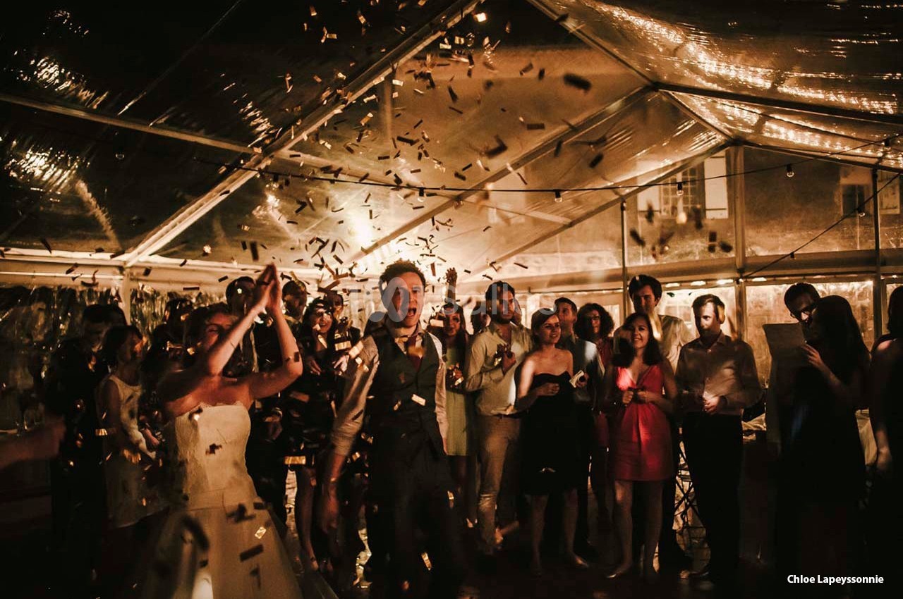 Des invités célèbrent sous une tente éclairée, entourés de confettis et d'une ambiance festive.