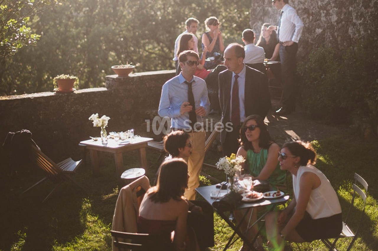 Groupe de personnes discutant autour de tables en extérieur, dans un jardin ensoleillé avec un mur en pierre.