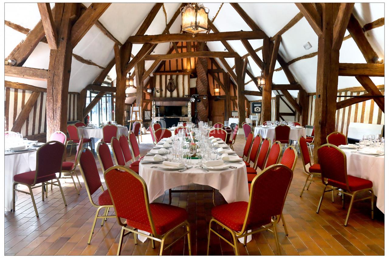 Salle de réception avec poutres apparentes, tables rondes dressées avec nappes blanches et chaises rouges.