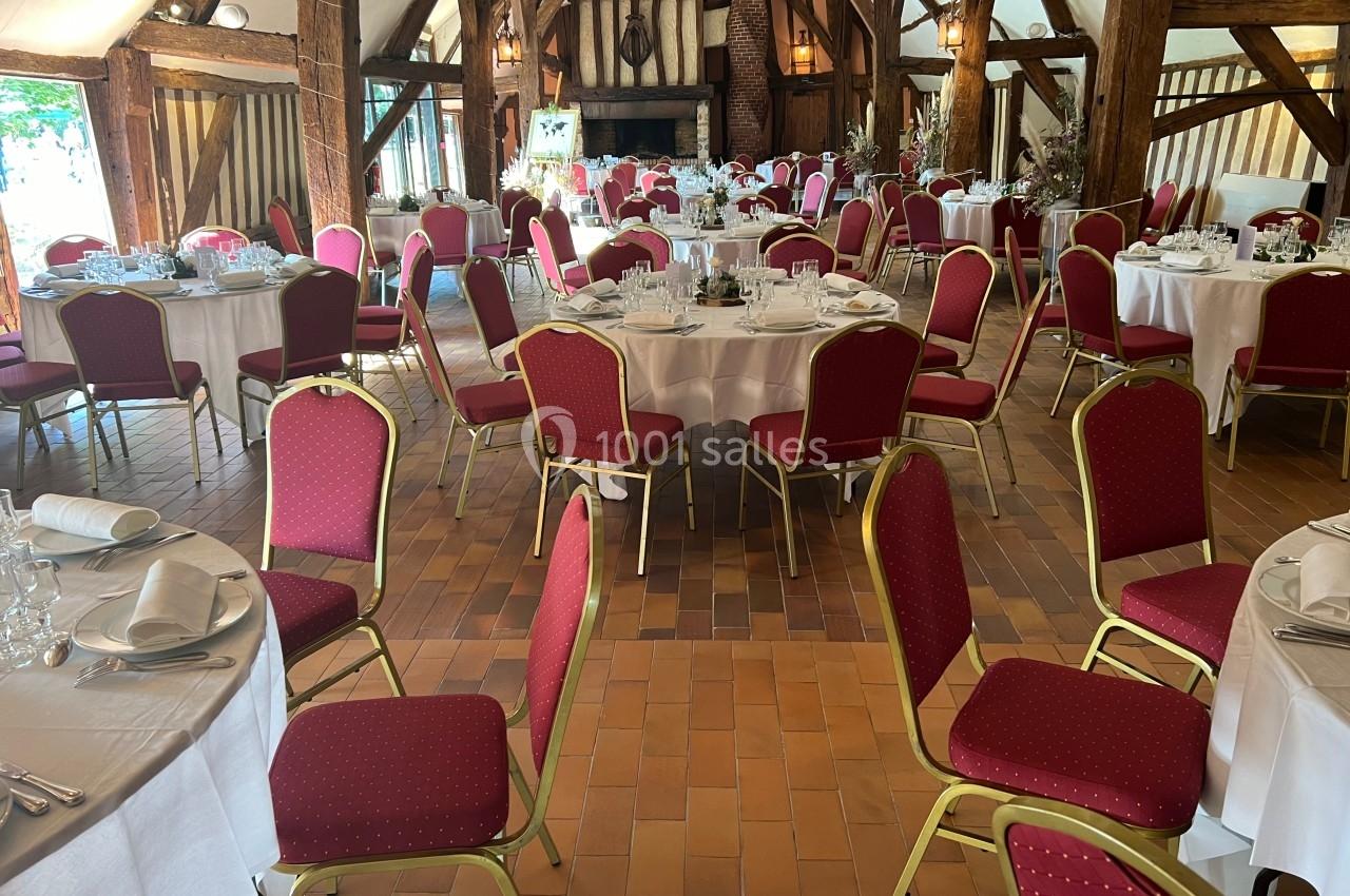 Salle de réception avec tables rondes dressées, nappes blanches et chaises rouges, dans un cadre rustique avec poutres…