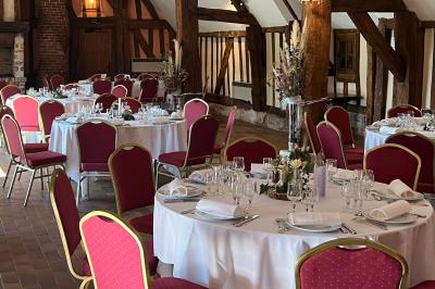 Salle de réception rustique avec poutres en bois apparentes, tables rondes dressées et chaises rouges.