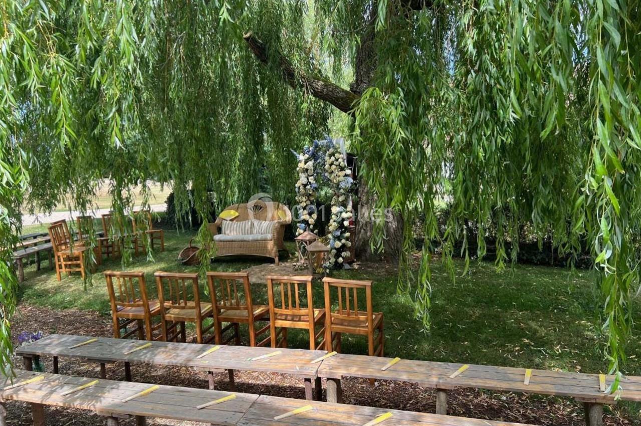 Chaises en bois disposées sous des arbres avec un banc décoré de fleurs pour une cérémonie en plein air.