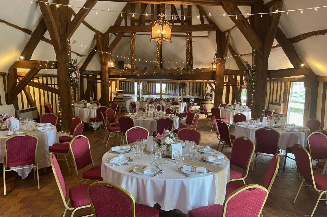 Salle de réception rustique avec poutres apparentes, tables rondes dressées et chaises rouges, décorée de guirlandes…