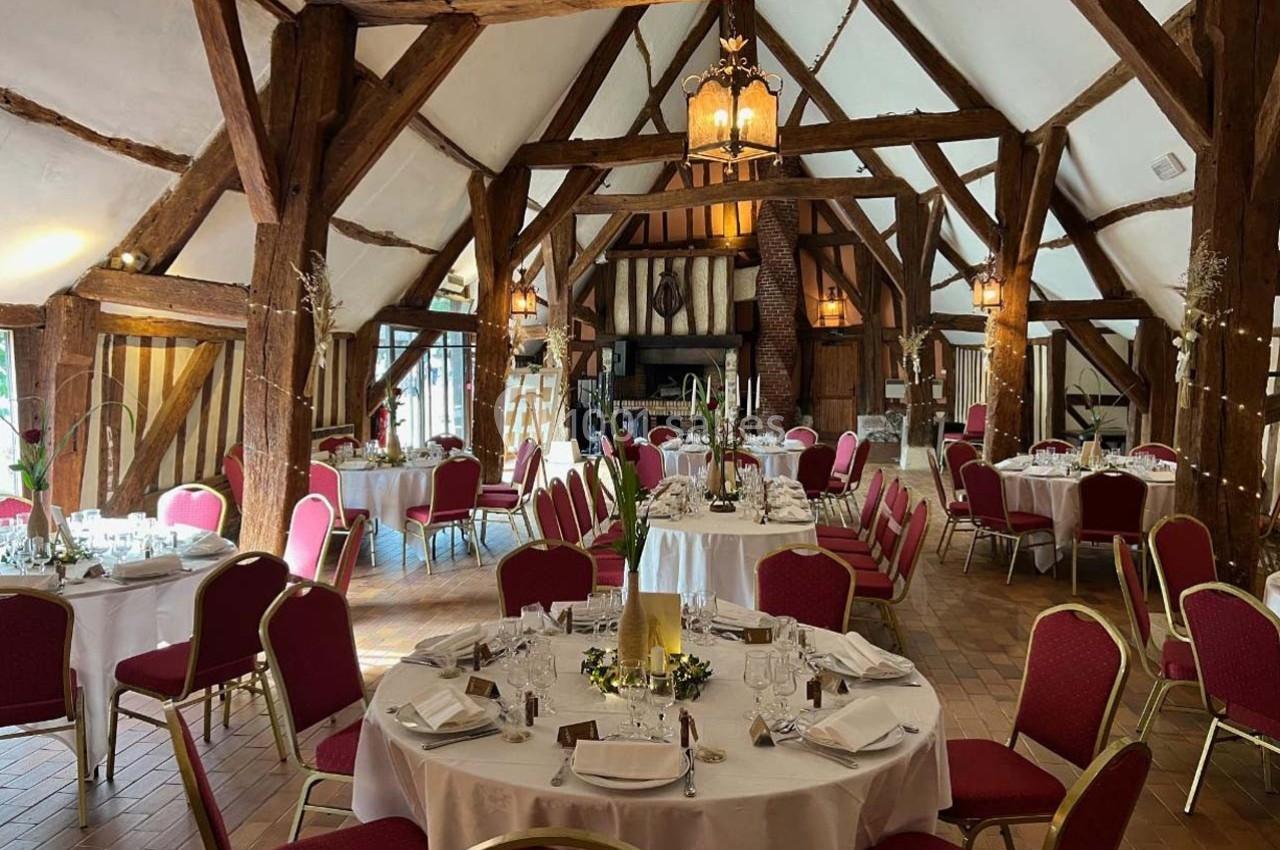 Salle de réception rustique avec poutres apparentes, tables rondes dressées et chaises rouges disposées pour un événement.