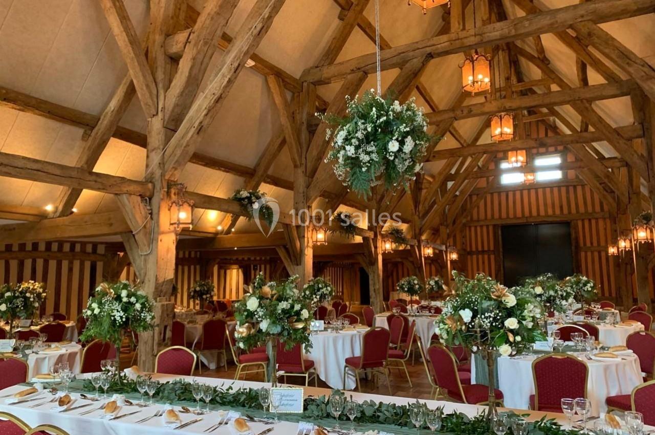 Salle de réception avec poutres apparentes, tables décorées de fleurs et chaises rouges disposées pour un événement.