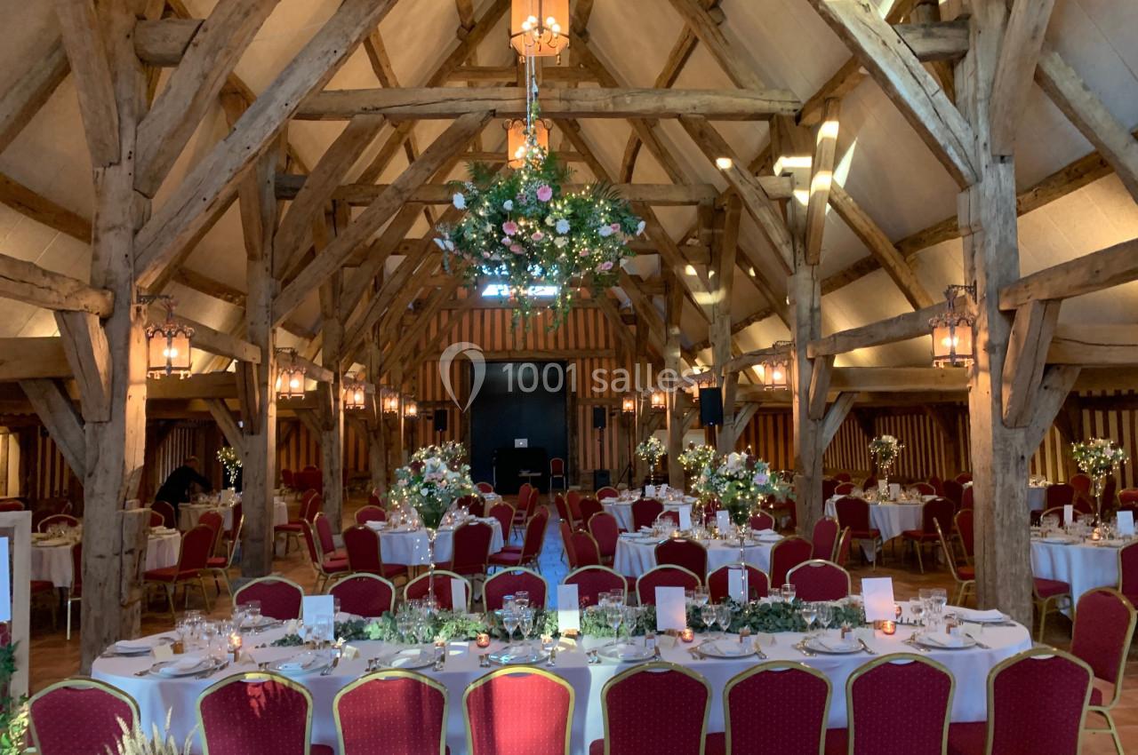 Salle de réception avec charpente en bois, tables décorées de fleurs et chaises rouges alignées.