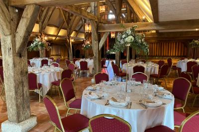 Salle de réception rustique avec poutres en bois apparentes, tables rondes dressées et chaises rouges.