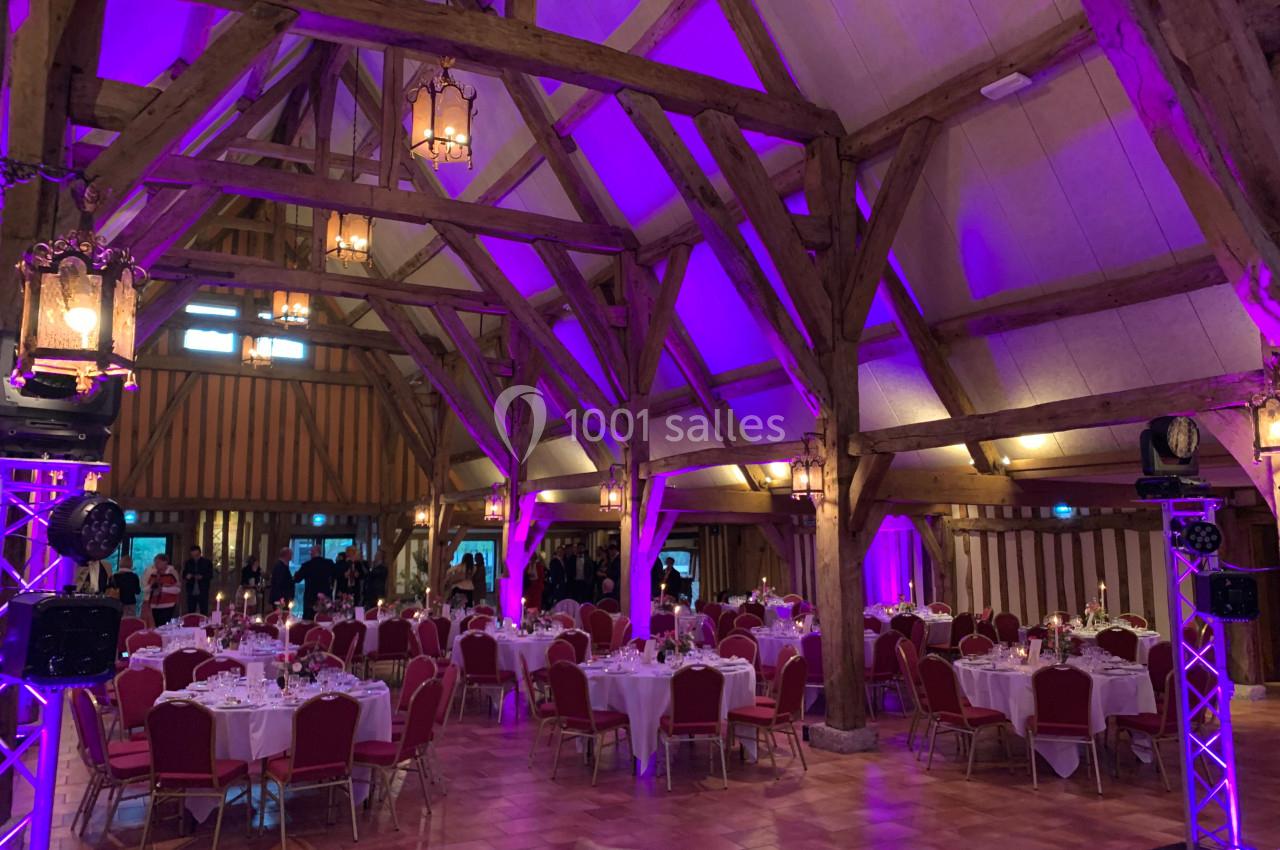Salle de réception avec charpente en bois, éclairage violet et tables rondes dressées pour un événement.