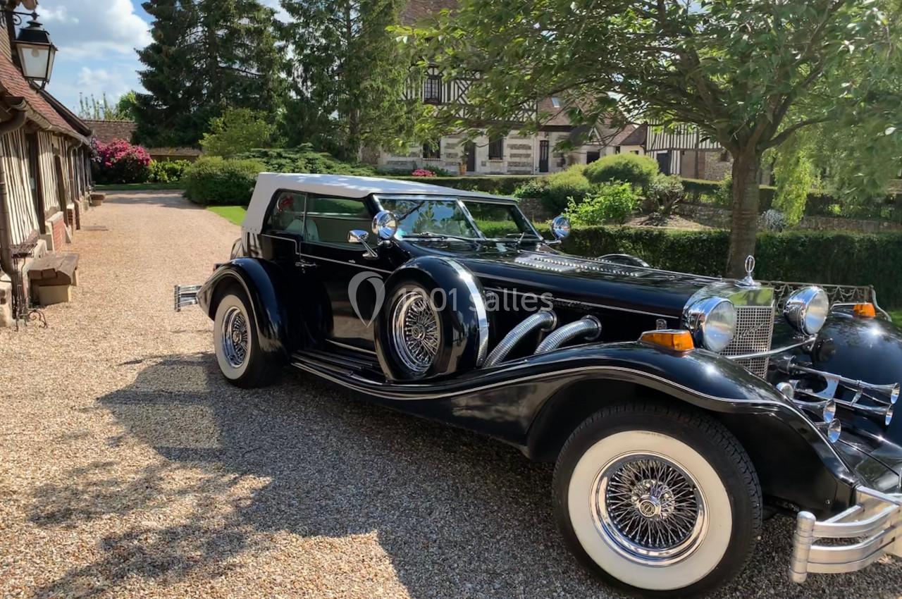Voiture ancienne noire avec toit blanc garée sur une allée en gravier, entourée de verdure et de bâtiments à colombages.