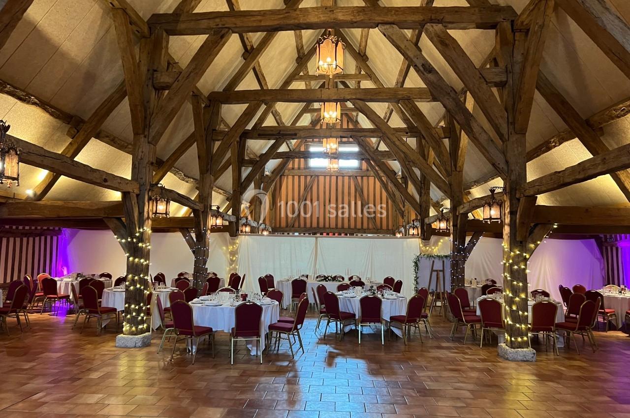 Salle de réception avec charpente en bois apparente, tables rondes dressées et chaises rouges sur un sol en carrelage.