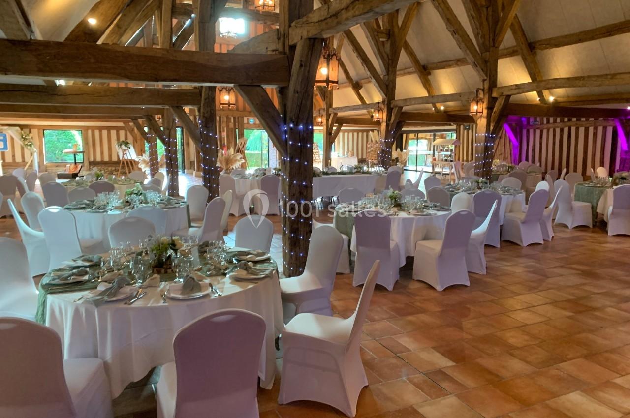 Salle de réception rustique avec poutres apparentes, tables rondes dressées et chaises blanches, éclairage chaleureux.