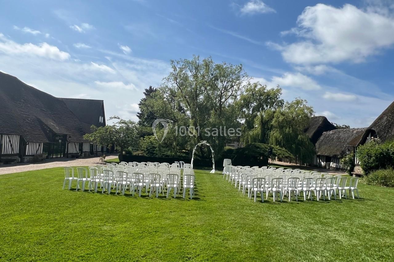 Chaises blanches disposées en extérieur sur une pelouse, formant une allée centrale, entourées de bâtiments à toit de chaume.