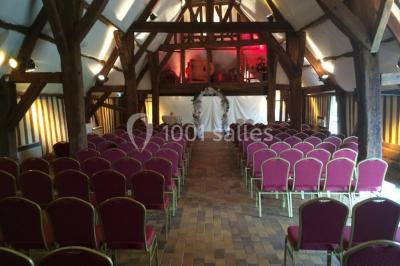 Salle de réception rustique avec poutres en bois apparentes, tables rondes dressées et chaises rouges.