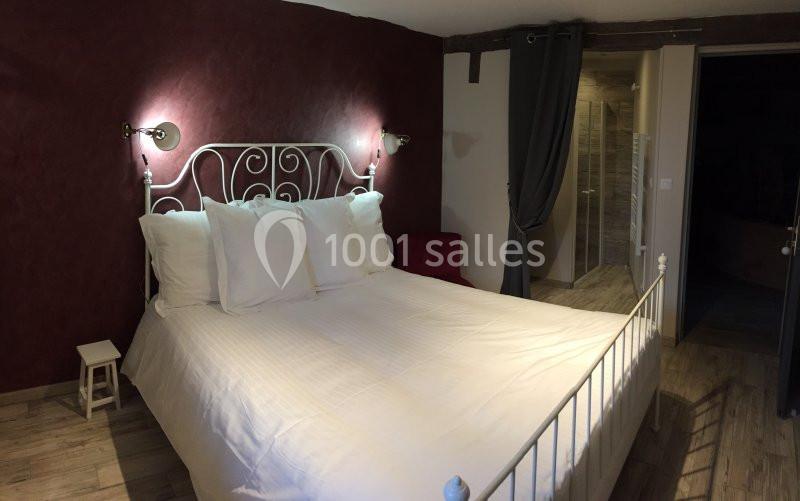 Lit double avec tête de lit en métal blanc, draps blancs, murs bordeaux et accès à une salle de bain attenante.