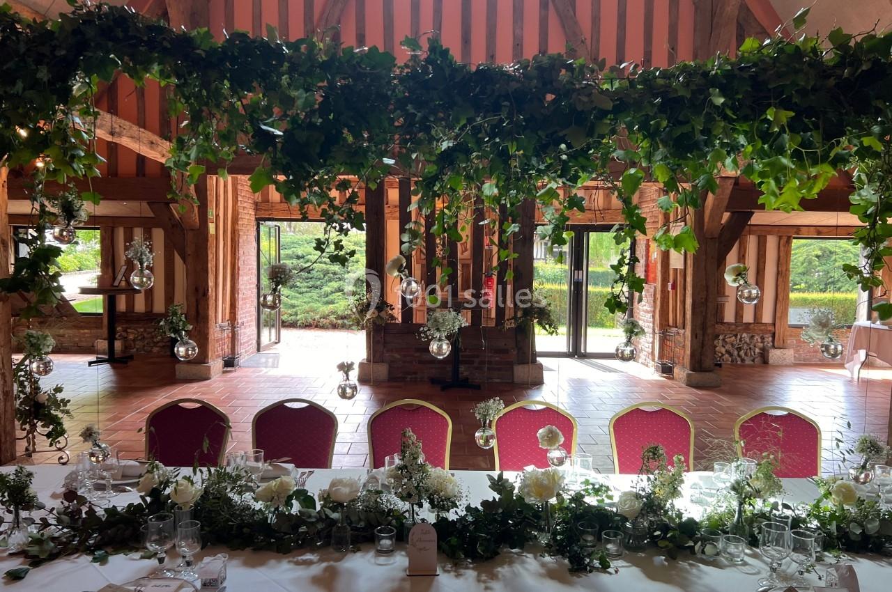 Salle de réception décorée avec des guirlandes de feuillage suspendues, des tables dressées et des chaises rouges.