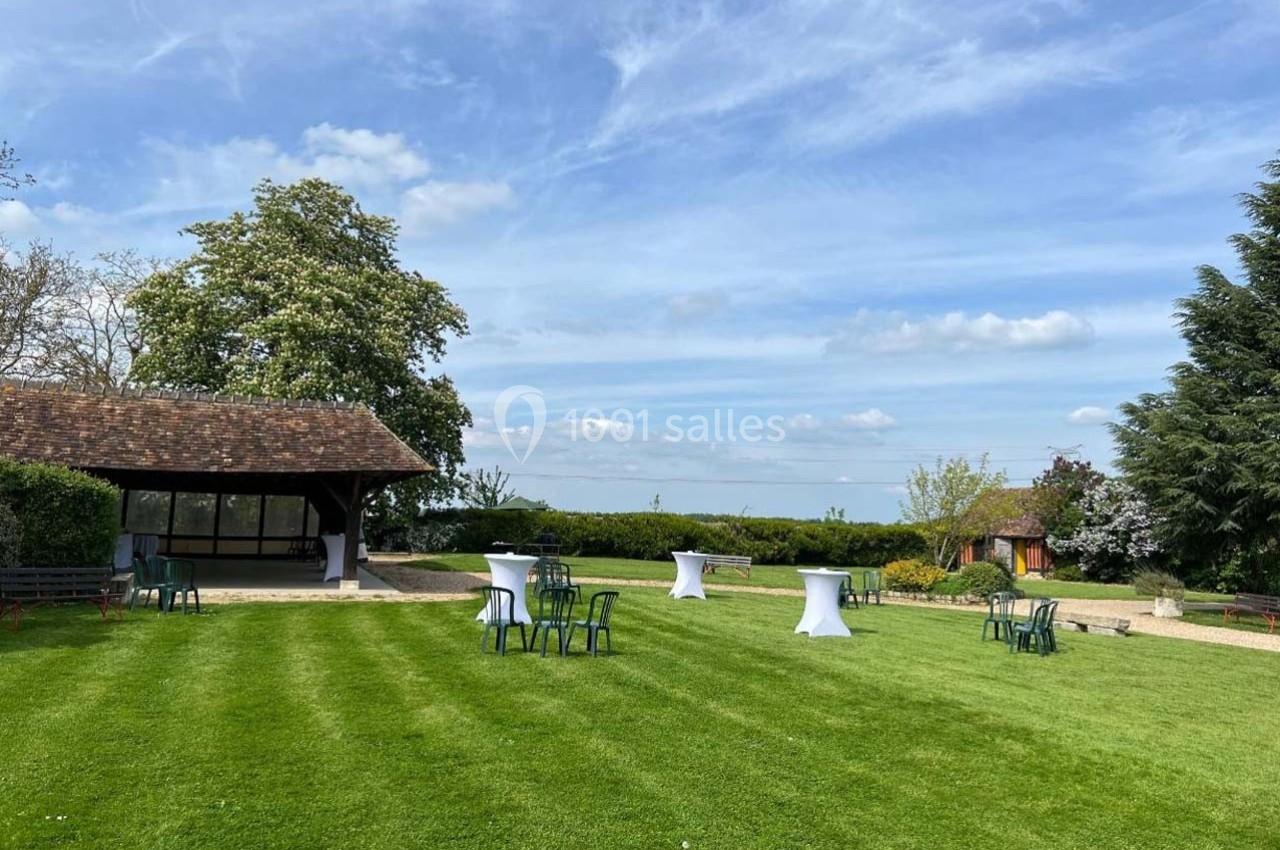 Pelouse verdoyante avec tables hautes et chaises, entourée d'arbres et d'une petite structure en bois sous un ciel dégagé.
