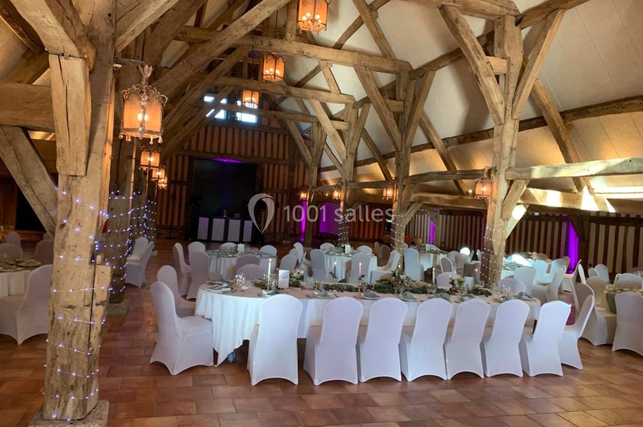 Grande salle de réception avec poutres apparentes, tables rondes dressées et chaises blanches, éclairage chaleureux.