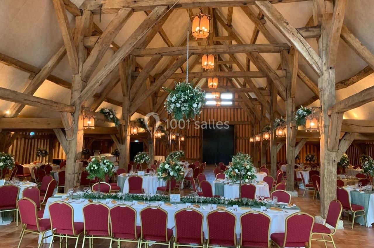 Salle de réception avec charpente en bois, tables dressées avec nappes blanches, chaises rouges et décorations florales.