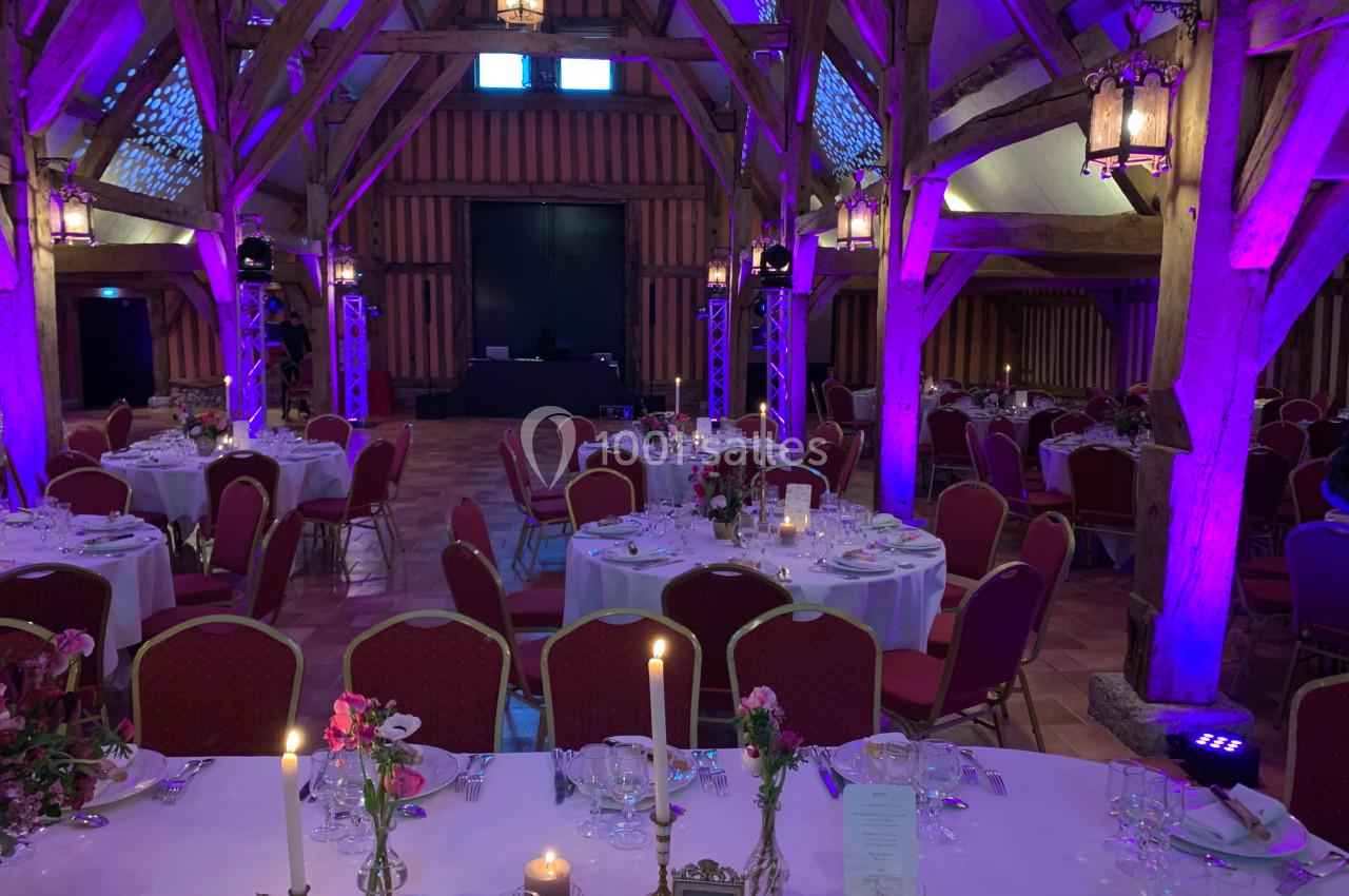 Salle de réception avec poutres apparentes, tables rondes dressées et éclairage violet.