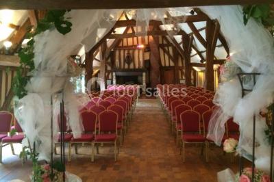Salle de réception rustique avec poutres en bois apparentes, tables rondes dressées et chaises rouges.