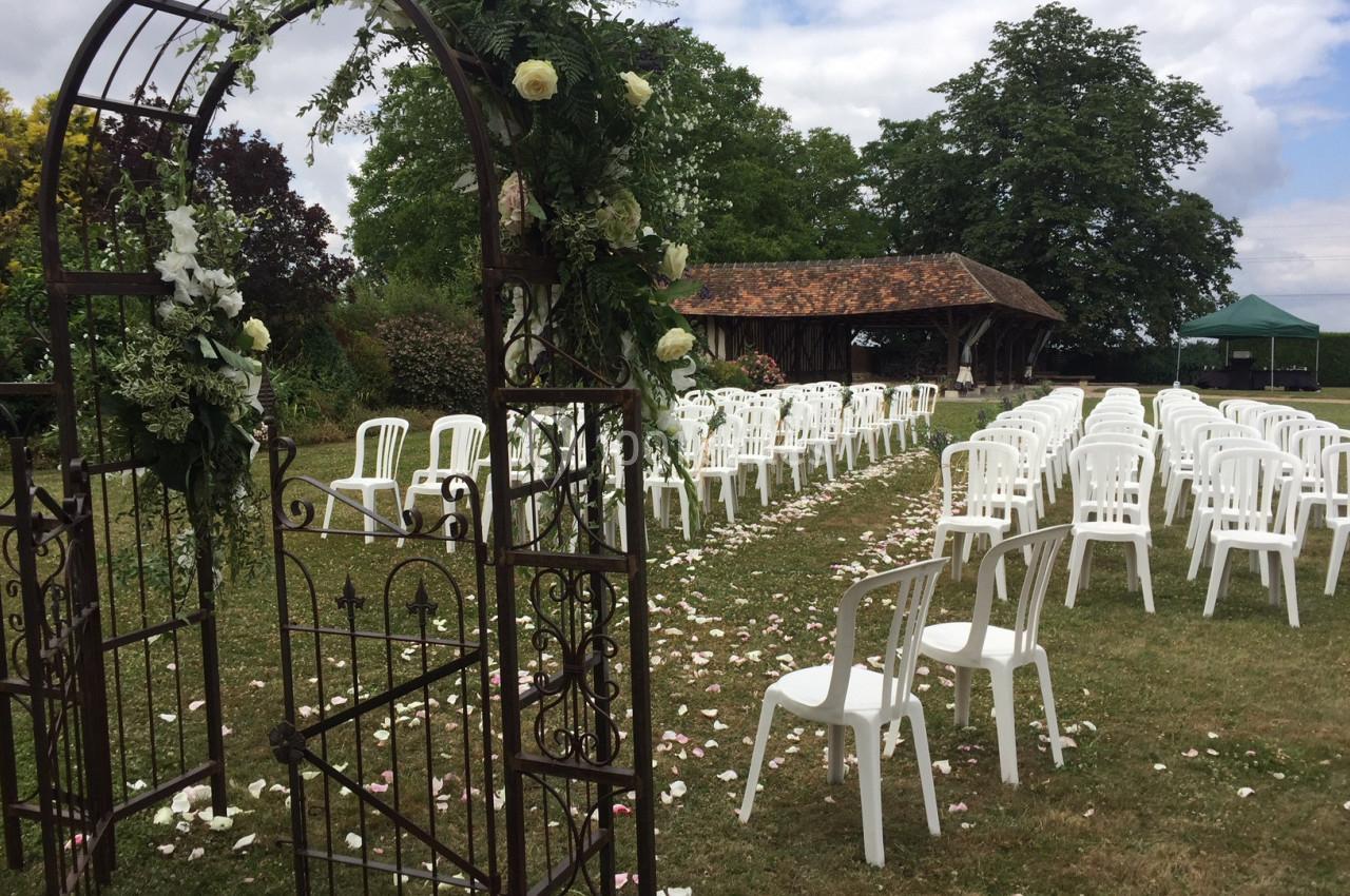 Chaises blanches alignées en extérieur pour une cérémonie, avec une arche fleurie et un chemin de pétales sur l'herbe.