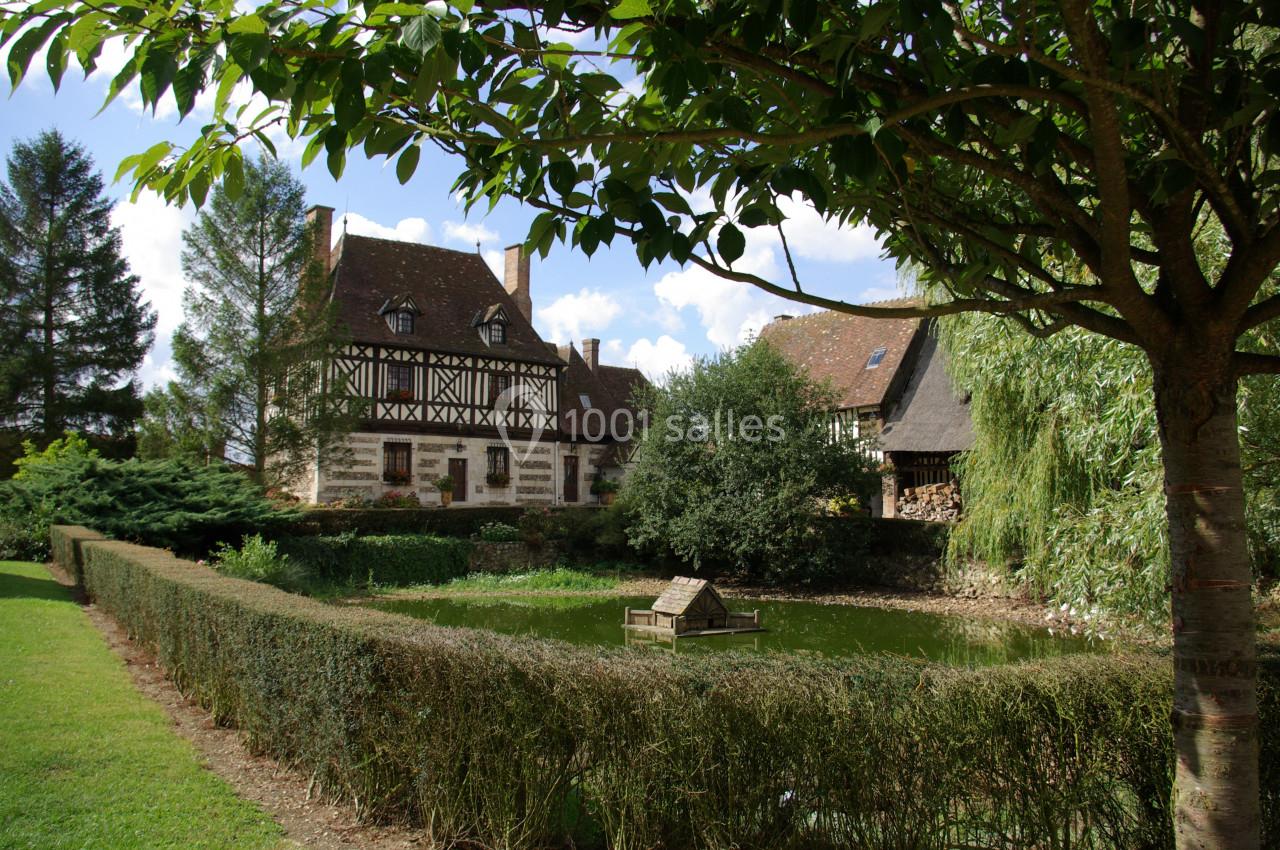 Maison à colombages entourée de verdure, avec un étang bordé de haies et un puits en bois au centre.