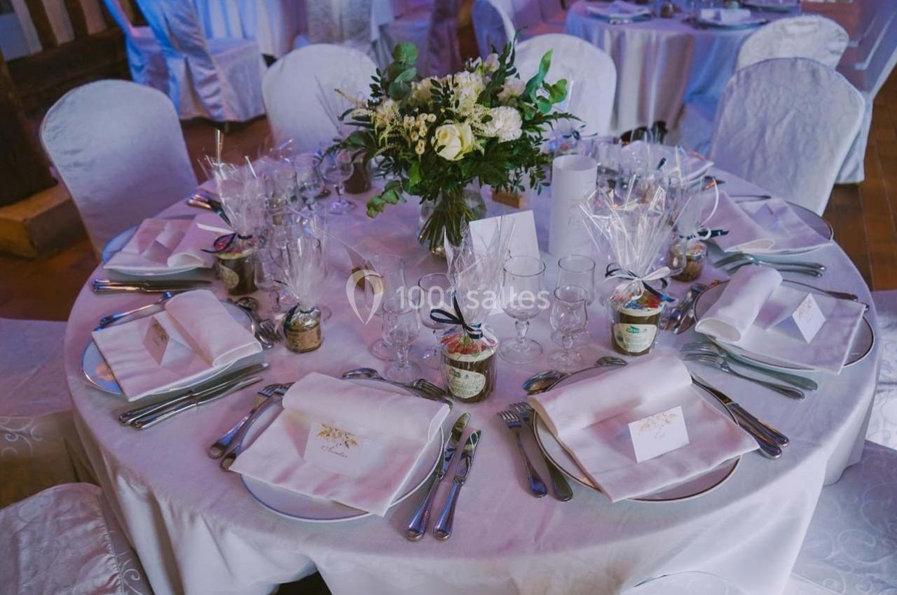 Table ronde élégamment dressée avec nappes blanches, couverts, verres, cadeaux invités et centre de table floral.