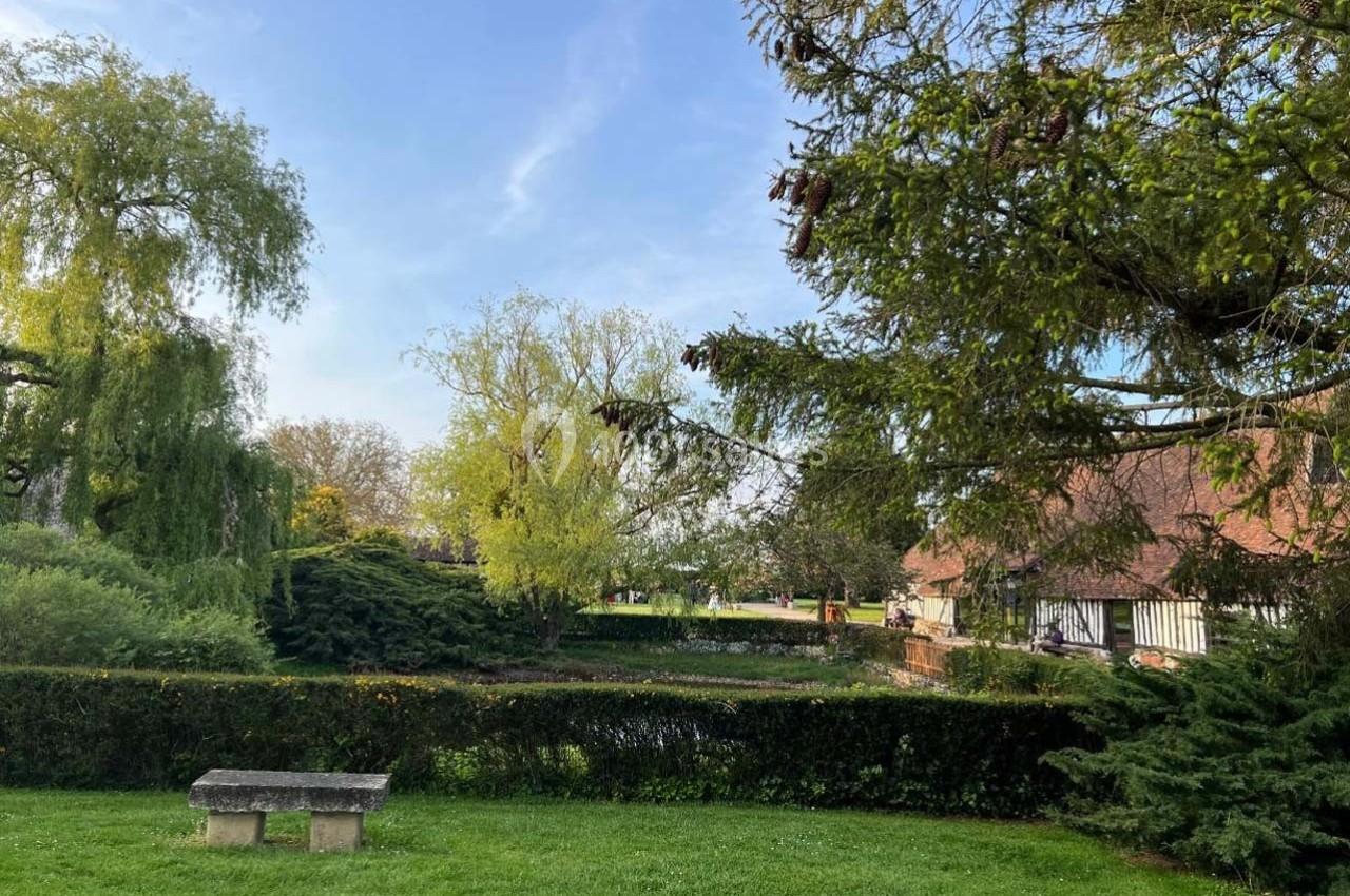 Un jardin verdoyant avec des arbres, une haie, un banc en pierre et une maison à colombages en arrière-plan.