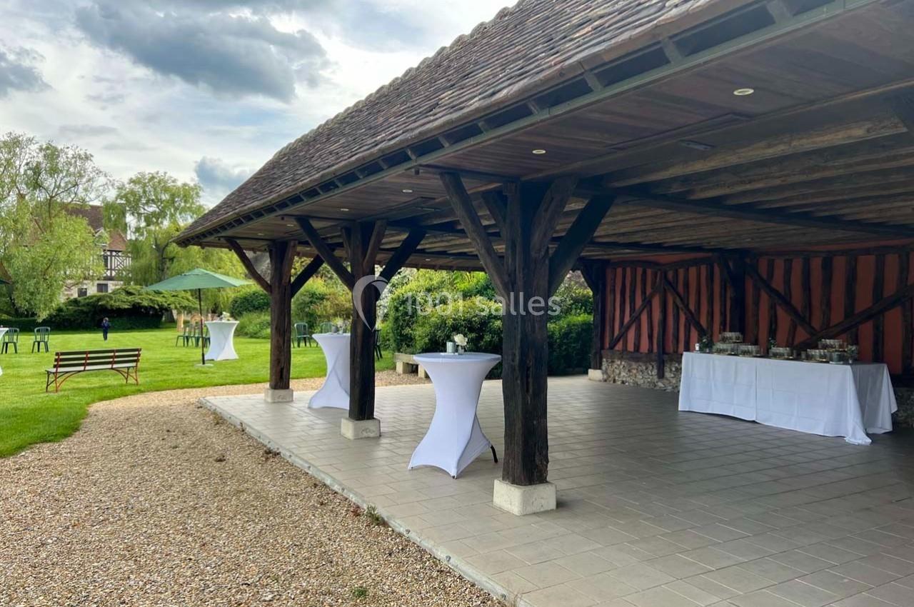 Abri en bois avec toit en tuiles, ouvert sur un jardin verdoyant, tables hautes et buffet installés pour un événement.